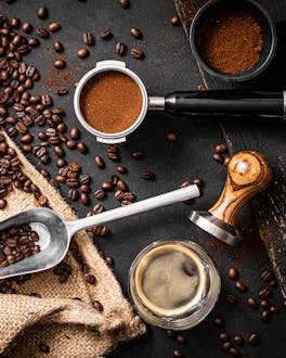 Zubehör und Zutaten für einen leckeren Espresso: Geröstete Kaffeebohnen, eine kleine Kaffeeschaufel, ein Kaffeesack, ein gefüllter Siebträger mit feinem Kaffeemehl, ein Tamper mit Holzgriff und zwei Tassen mit frisch aufgebrühtem Espresso vor dunklem Hintergrund