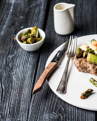 Steak mit Garnelen, grünem Spargel und Sauce Hollandaise auf weißem Teller mit Besteck auf dunklem Holzuntergrund.