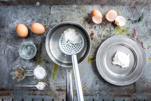 Ein Ei wird in einem Stahltopf pochiert, im Topf liegt eine Schöpfkelle. Neben dem Topf steht ein Metallteller mit einem bereits pochierten Ei. Es liegen Eier und Eierschalen sowie Löffel und Salz und Pfeffer im Bild