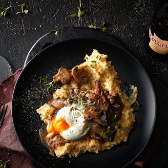 Ein schwarzer Teller mit cremiger Parmesan-Polenta, in Bier sautiertenPilzen und einem pochierten Ei neben einem Glas mit Duckstein
