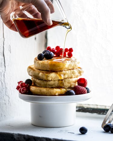 Fluffige Pancakes sind auf einem Podest aus Schüssel und Teller übereinandergestapelt und dekoriert mit gemischten Beeren. Gerade wird Ahornsirup aus einer Glaskaraffe darüber gegossen.