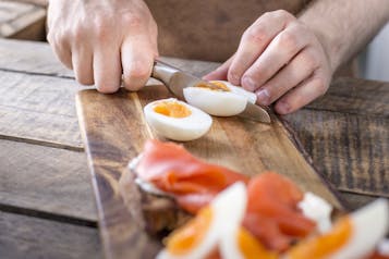 Auf einem Holzbrett wird ein gekochtes Ei mit einem Messer geschnitten.Unscharf im unteren Bildrand ein Brot mit Räucherlachs und Ei