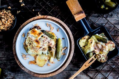 In einer Schale überbackene Cornichons, daneben ein Raclettepfännchen mit dem gleichen Gericht, das auf einem schwarzen Küchengitter steht