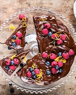 Angeschnittener Schokoladen-Heidelbeerkuchen auf einer Servierplatte und nebenliegend Besteck auf Holzuntergrund.