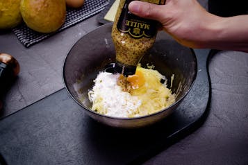 Aus einer Flasche wird Senf in eine Schüssel zu geriebenen Kartoffeln, Mehl und Ei gegeben.