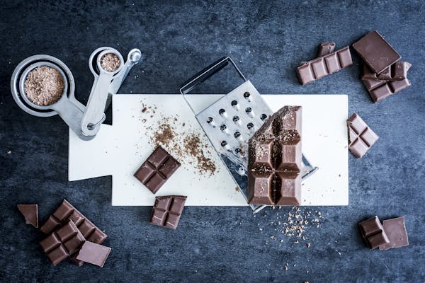 Topshot von oben auf ein weißes Schneidebrett, auf dem mehrere Tafelstücke von dunkler Schokolade und geraspelte Schokolade neben einer Küchenreibe auf dunklem Untergrund liegen