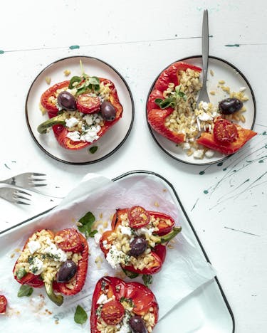 Rote Paprikahälften gefüllt mit Ebli, Tomaten, Oliven und Feta auf weißem Tablett und zwei weißen Tellern angerichtet