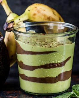 Grün-braun geschichteter Smoothie in Weckglas neben Avocado und zwei Bananen