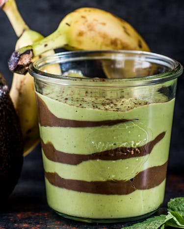 Grün-braun geschichteter Smoothie in Weckglas neben Avocado und zwei Bananen