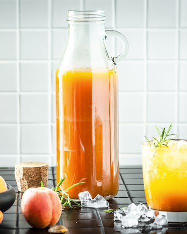 Eine große Flasche, gefüllt mit Aprikosensirup steht vor weißen Kacheln. Daneben ein Glas mit Sirup-Gemisch und einige Aprikosen liegen in einer Schale daneben.