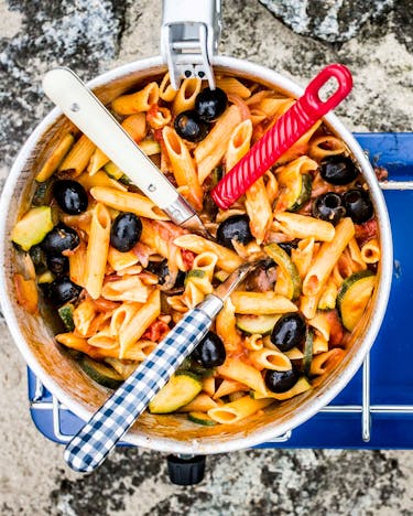 Drei Gabeln in Topf mit Nudeln in Tomatensauce mit Oliven und Zucchini auf blauem Gaskochfeld von oben