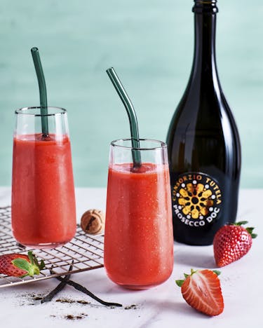 Zwei Cocktailgläser mit eisgekühltem Erdbeer Prosecco Slush auf einem hellen Untergrund neben frischen Erdbeeren und Vanilleschoten vor türkisem Hintergrund. Dazu eine Flasche Prosecco DOC.
