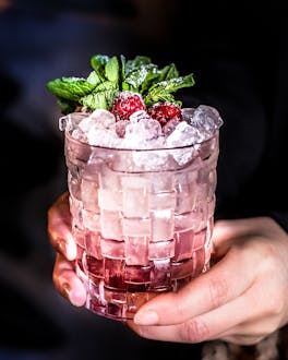 Eine Hand hält ein Glas Raspberry Cocktail, garniert mit Minze und Himbeeren