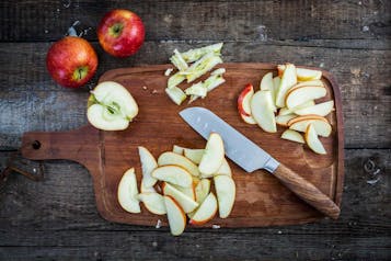Auf einem Holzbrett liegen Apfelscheiben, ein halber Apfel und etwas Küchenabfall. Dazwischen ein großes Küchenmesser. Neben dem Brett liegen zwei ganze Äpfel