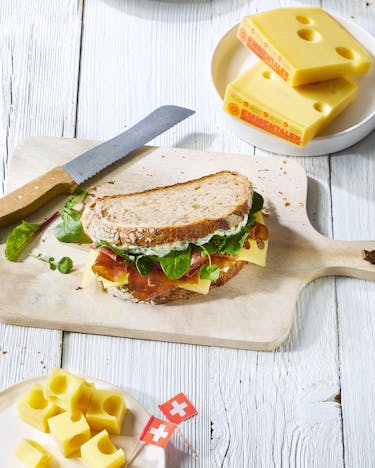 Ein Sandwich liegt auf einem Holzbrett. Daneben liegen ein Brotmesser und ein paar Stücke Emmentaler auf einem weißen Teller.