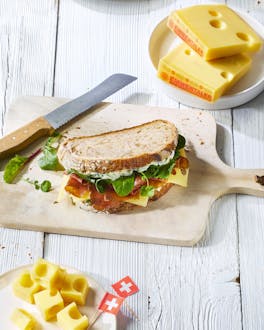 Ein Sandwich liegt auf einem Holzbrett. Daneben liegen ein Brotmesser und ein paar Stücke Emmentaler auf einem weißen Teller.