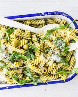 Weiße Auflaufform mit blauem Rand auf weißem Tisch gefüllt mit Fusilli, Pesto, Rucola und Oliven