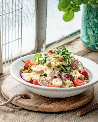 Nudelsalat mit Mozzarella, Thunfisch und anderen Zutaten in weißem Teller vor einem hellen Fenster