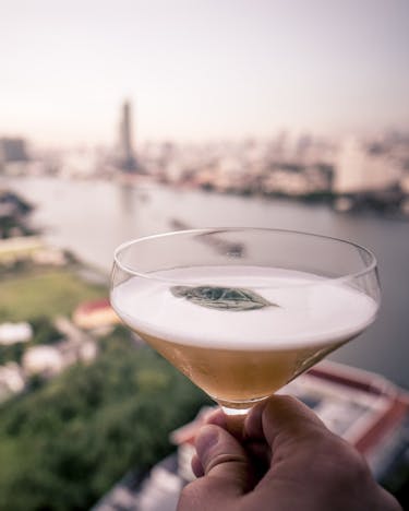 Ein Drink wird hochgehalten. Im Hintergrund die Skyline von Bangkok.
