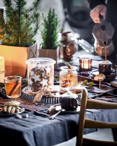 Adventskaffeeklatsch-Szenerie am Tisch, der Tisch ist weihnachtlich gedeckt, Lichter, Bäume und Kerzen stehen überall, Plätzchen in Dosen sind zu sehen. Im Hintergrund wird Kaffee eingegossen.