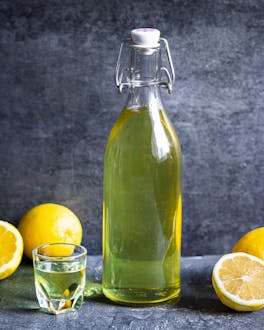 Limoncello in einer Glasflasche vor dunklem Hintergrund mit Zitronen und einem Shotglas.