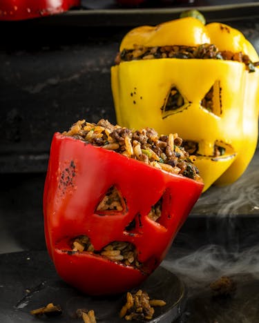 Zwei gelbe und eine rote gefüllte Paprika mit eingeritzten Gesichtern auf einem schwarzen rauchenden Hintergrund