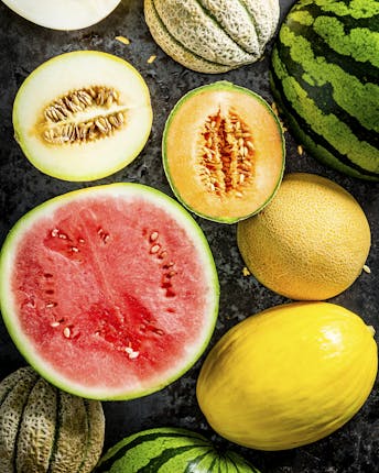 Buntes Allerlei verschiedener Melonenarten, teils ganz, teils halbiert, als Topshot vor dunkelgrauem Hintergrund