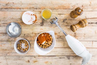 Kichererbsen mit Crème Fraîche und Harissapaste in einem hohen Gefäß, daneben Schüsseln der einzelnen Komponente, ein Pürierstab und Salz- und Pfeffermühlen auf einem hellen Holzhintergrund