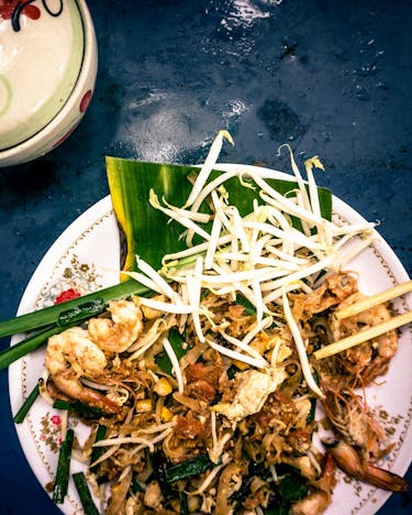 Pad Thai mit Garnelen, Tofu und Sojasprossen auf einem weißen Teller angerichtet, an dem zwei Essstäbchen lehnen. Im oberen linken Bildrand eine kleine bunte Schale mit Deckel.