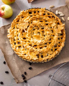 Flecht-Pie mit Apfel und Amaretto auf einem braunen Tuch. Daneben liegen Äpfel und ein Nudelholz.