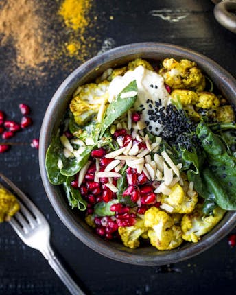 Curry-Zimt-Blumenkohl, Spinat, Granatapfelkerne und Mandelsplitter in einer Schüssel auf einem dunklen Untergrund auf dem Currypulver verteilt liegt.