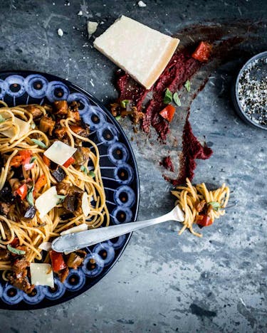 Spaghetti mit Auberginen-Paprika-Pesto auf eine blauen Teller, daneben ein Stück Parmesan.