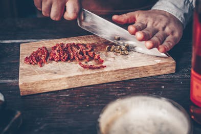 getrocknete Tomaten in Streifen geschnitten auf einem Holzbrett, daneben werden Kapern mit einem großen Messer gehackt