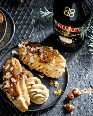 Eclairs gefüllt mit Espressocreme, glasiert mit Baileys und Haselnüssen garniert auf dunklen Desserttellern serviert neben einer Baileys Flasche.
