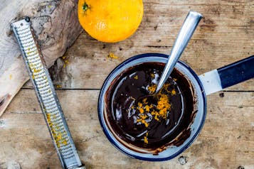 geschmolzene Schokolade in einem kleinen Topf, darin Orangenabrieb, daneben eine Orange und eine kleine Reibe