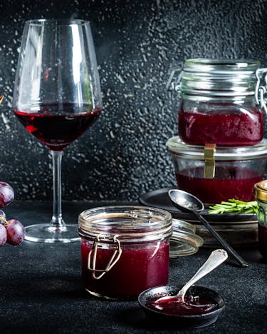 vor schwarzem Hintergrund stehen mehrere Gläser mit Trauben-Konfitüre und ein Glas Rotwein