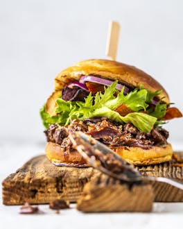 Auf einem Holzbrett vor weißem Hintergrund liegt in Burger mit Jackfruit, Salat und Zwiebelringen. Davor lehnt ein Esslöffel.