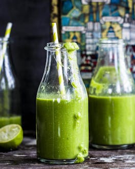Drei kleine Glasflaschen mit grünem Smoothie und Strohalmen auf dunklem Holzgrund