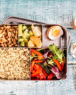 Couscous mit Gemüse, einem Ei und Erdnüssen in einer Bento-Box mit verschiedenen Fächern auf grünlichem Holzuntergrund