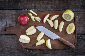 Auf einem Holzbrett liegen teilweise geschnittene Äpfel, außerdem eine aufgeschnittene Zitrone und ein Messer