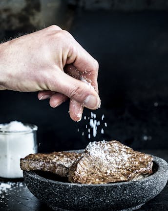 Gebratene Fleischstücke in einer dunklen Schale neben einem Salzglas. Eine tattowierte Hand streut grobe Salzkörner auf das Fleisch.