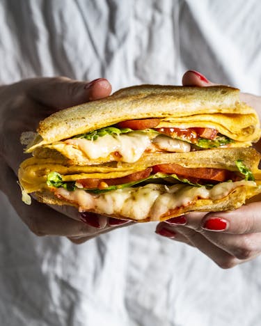 Omelettsandwich mit Tomate und Chorizo in Händen, von vorne im Anschnitt fotografiert, der Käse läuft etwas heraus.