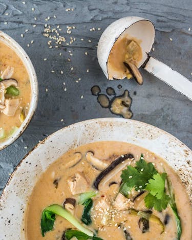 Asiatische Suppe mit Kokos, Shiitage und Huhn in Suppenschale, nebenliegend eine Suppenkelle auf grauem Untergrund.