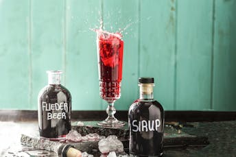 Aus einem hohen Glas spritzt Fliederbeersirup heraus. Davor stehen zwei selbstbeschriftete Flaschen gefüllt mit rotem Sirup.