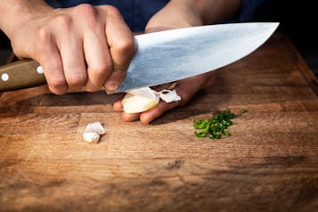 Knoblauch wird mit einem großen Messer geschält, auf dem Holzbrett darunter liegt Knoblauchschale und geschnittene Chili