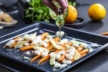 Möhren, Zwiebeln und Ingwer liegen auf einem Backblech un werden mit Olivenöl begossen.