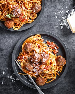 Schwarze Teller mit Spaghetti, Tomatensauce, Fleischbällchen und extra Teller mit Parmesan und Basilikum.