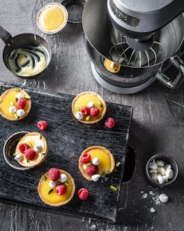 Mehrere Zitronen-Himbeer-Tartelettes auf schwarzem Brett und Krups Standmixer.