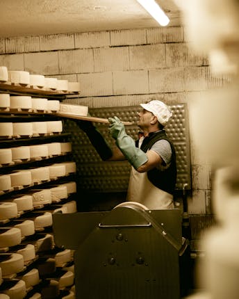 Ein Käser gibt ein frisches Holzbrett mit einem runden Käselaib zur Lagerung neben die vielen anderen aufgetürmten Käse