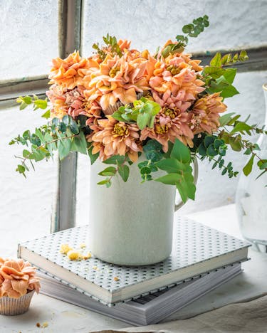 Ein Strauß mit aprikotfarbenen Chrysanthemen und identisch dekorierten Blumen-Cupcakes steht als Bouquet in einer weißen Vase auf der Fensterbank. Daneben stehen Sektgläser zum Anstoßen bereit.